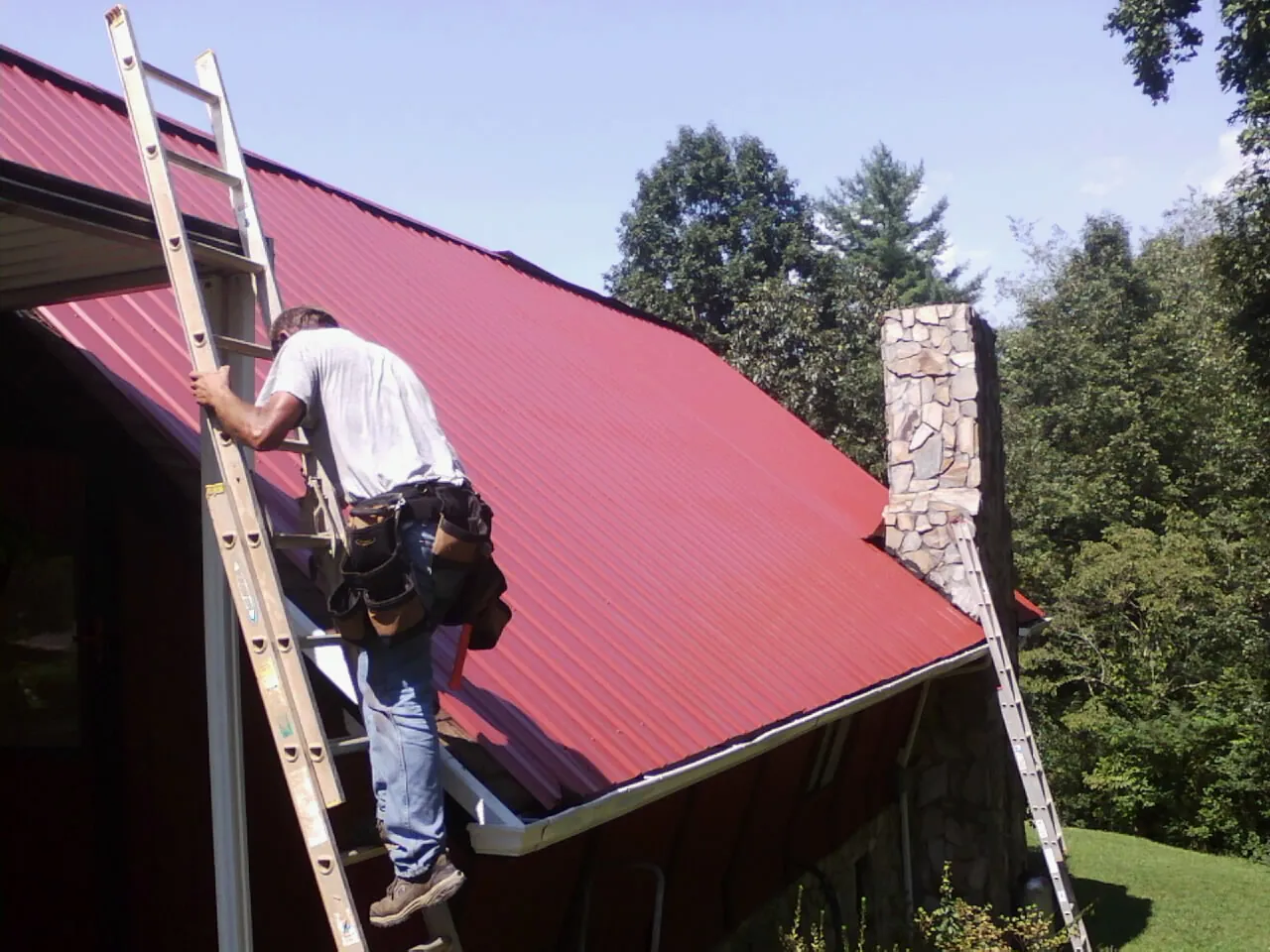 Roofer on a ladder beside a red metal roof and stone chimney — Soffit & Fascia Repair by Roofing Brothers in Boonville